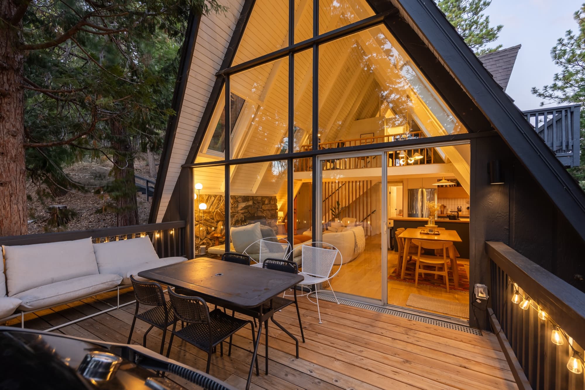 A-frame exterior at twilight with full glass wall glowing warmly, deck furniture, and pine trees