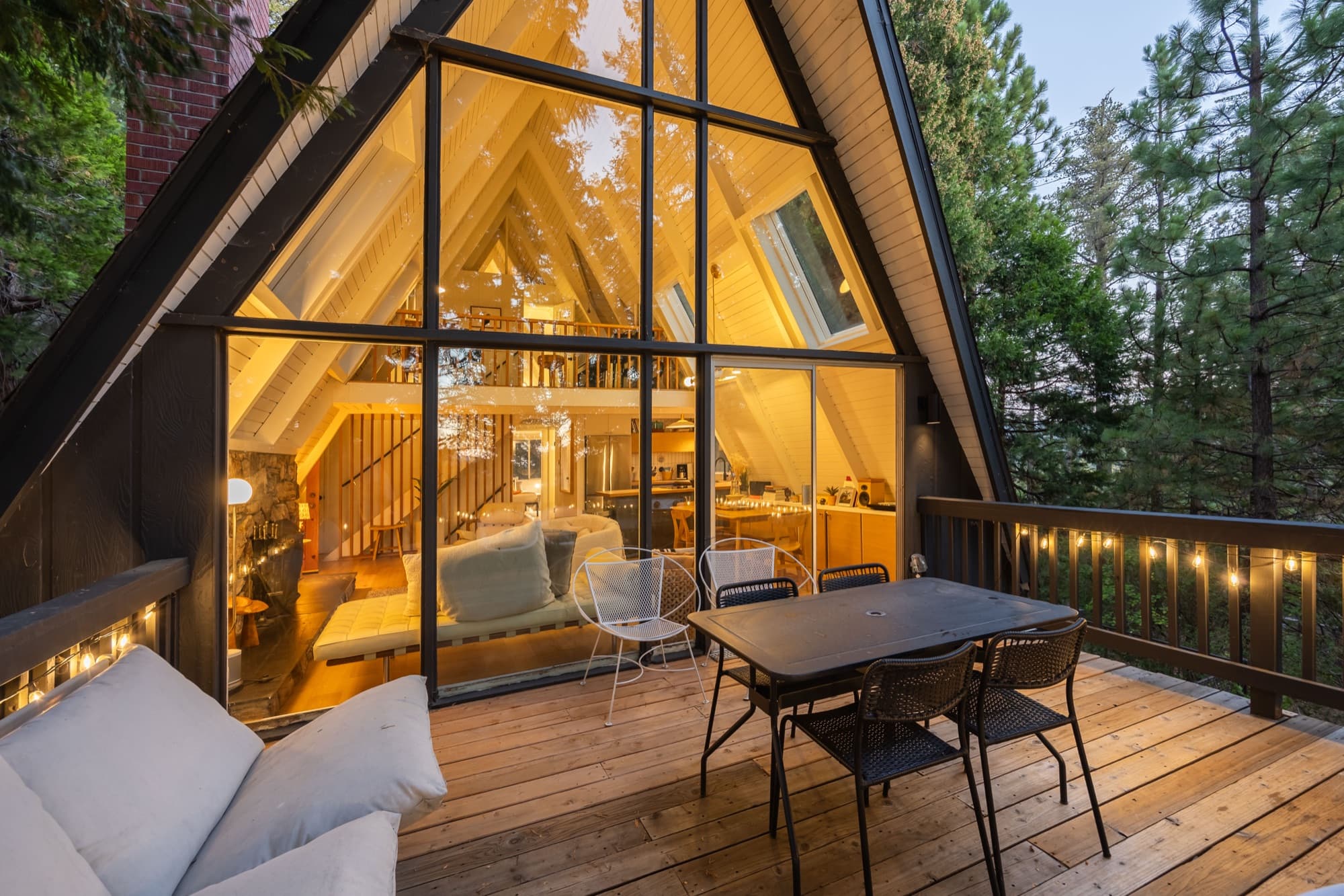 A-frame exterior from deck showing full glass facade, warm interior glow, and pine trees at dusk